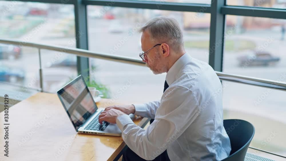Side view of businessman working on a computer. Mature man is typing on the laptop in the office on the city background through the window. Side view of businessman working on a computer. Mature man is typing on the laptop in the office on the city background through the window.