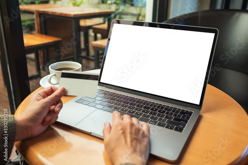 Mockup image of a woman holding credit card while using laptop with blank white screen and coffee cup on wooden table