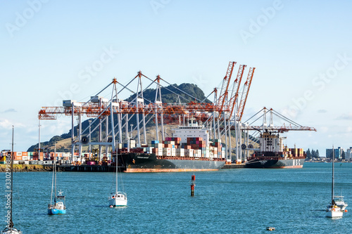 TAURANGA, NEW ZEALAND - MARCH 6, 2020: Cargo ships docked into Tauranga Harbour Port waiting for the adjacent container cranes to load. Mount Maunganui in the background.