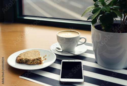 Cup of coffee, cookie and smartpohne with empty screen on a wooden table with stripes background near the window with copy space. Coffee shop and relax.
