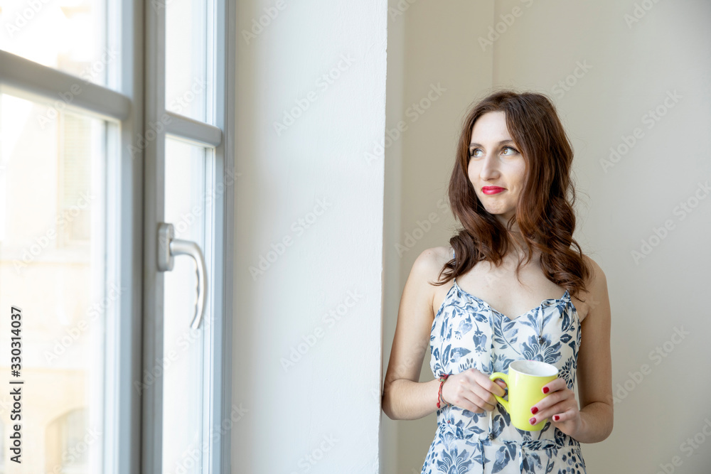 Brunette girl with long hair looks out the window with a yellow cup of tea.