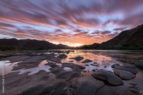 A beautiful long exposure landscape taken after sunset with mountains and the Orange River, with dramatic orange moving clouds reflecting in the water’s surface, taken in the Richtersveld South Africa