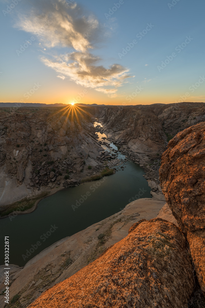 Obraz premium A beautiful vertical landscape view of the Augrabies Falls Gorge, mountains and river in South Africa, taken at sunset with a sunburst on the horizon and clouds in the blue sky.