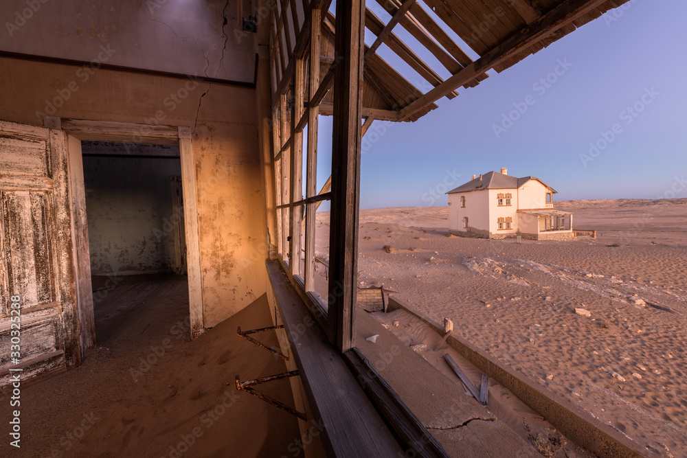 A photograph inside an abandoned house after sunset, with desert sand and a house visible through broken windows, taken in the ghost town of Kolmanskop, Namibia.