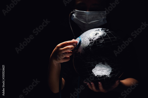 Hands of kid hold globe put stethoscope on sphere listen heartbeat, face covered in mask on black background. Ecological problems disasters and COVID-19 2019 2020 pandemic infection disease concept