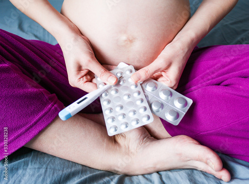 A young pregnant woman holds blisters with pills and a thermometer in front of her.