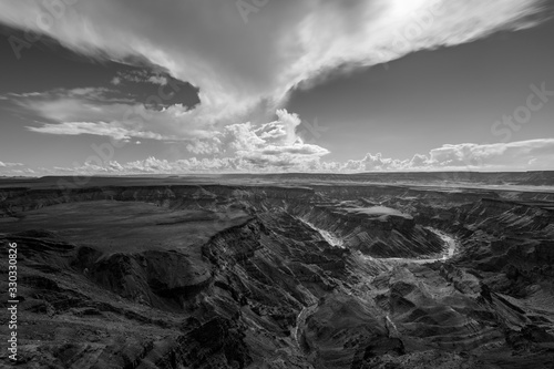 A black and white landscape taken at sunset on a stormy day on top of the arid and stark Fish River Canyon, Namibia, with the gorge and river in the foreground.