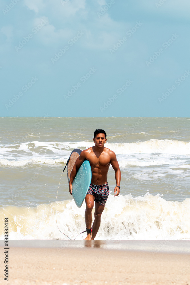 Young handsome man wet all over body holding surfboard with right hand,walking on the beach,show fit and firm body,model posing,blurry light around Young handsome man wet all over body holding surfboard with right hand,walking on the beach,show fit and firm body,model posing,blurry light around
