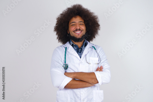 Brunette doctor man with happy face smiling with crossed arms looking confident at the camera standing against gray wall. Positive person.