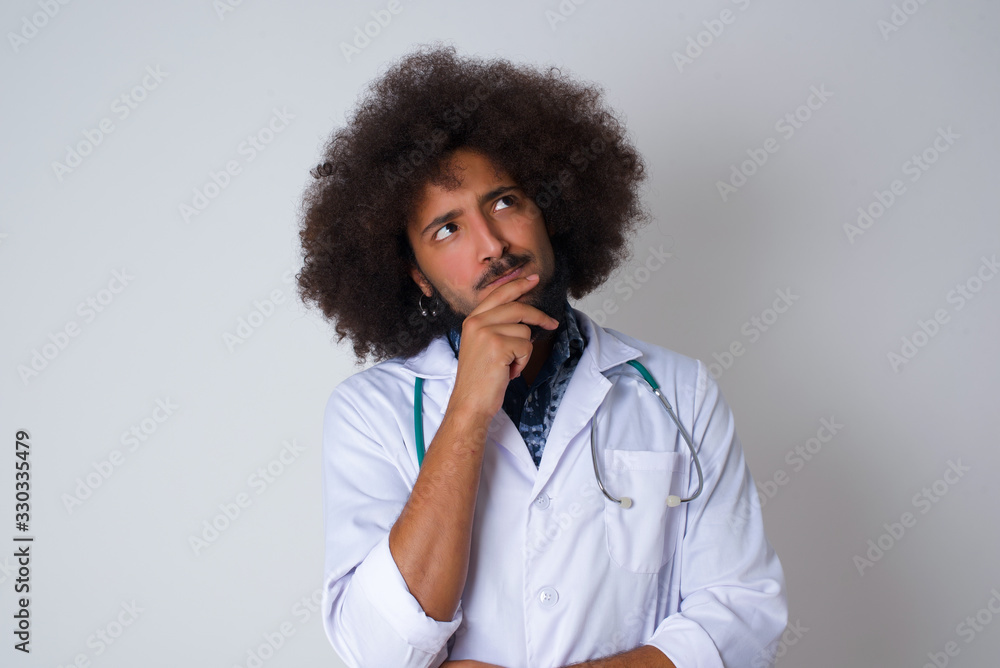 Shot of contemplative thoughtful Young African American doctor man keeps hand under chin, looks thoughtfully upwards, wearing medical uniform, poses over white background with free space for your text Shot of contemplative thoughtful Young African American doctor man keeps hand under chin, looks thoughtfully upwards, wearing medical uniform, poses over white background with free space for your text