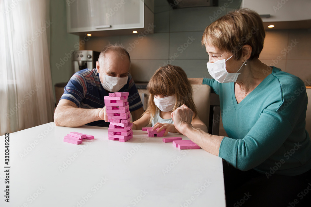 grandparents with granddaughter wearing medical face mask playing  board game together at home. quarantine. health concept. Corona Virus.  - obrazy, fototapety, plakaty 