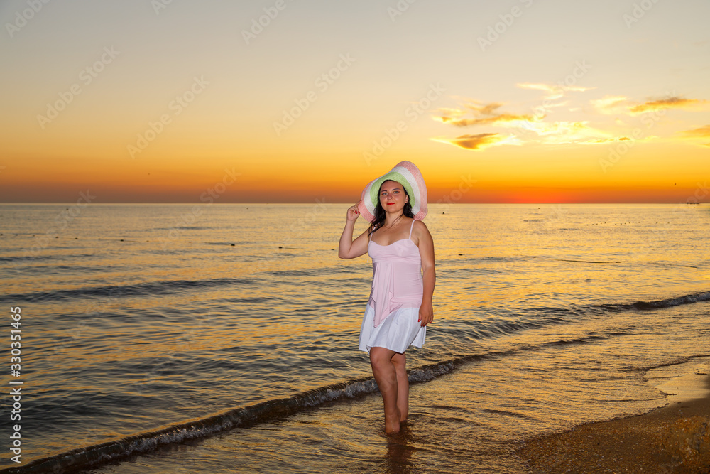 A young woman in a white dress and hat stands on the seashore at sunset. A young woman in a white dress and hat stands on the seashore at sunset.