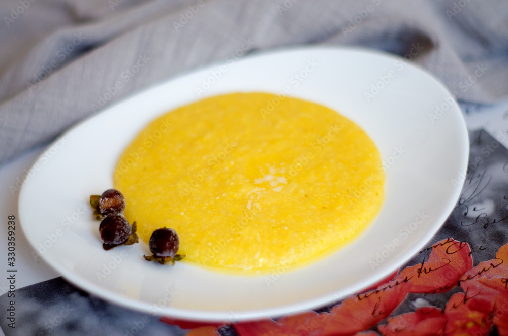 A portion of corn porridge with a piece of butter on a white plate decorated with frozen berries. C lose- up, selective focus, blur. A portion of corn porridge with a piece of butter on a white plate decorated with frozen berries. C lose- up, selective focus, blur.