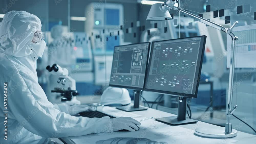 Electronics Factory Cleanroom: Engineer / Scientist in Coveralls Wors on Computer, Screen Shows Infographics and Software System Control UI, Developing Electronics for Medical Electronics