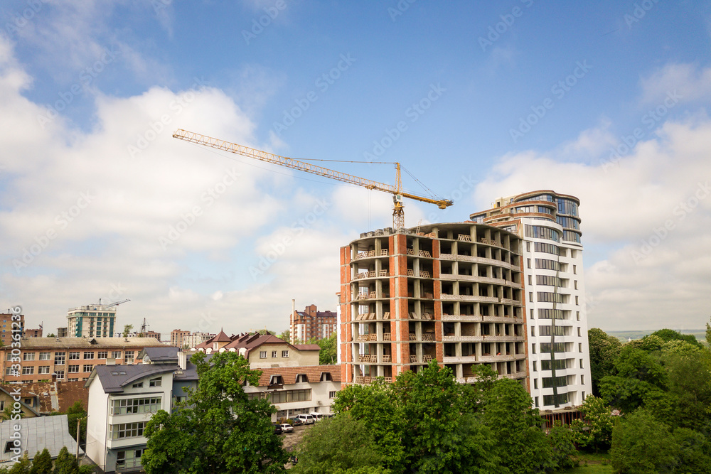 Apartment or office tall building unfinished under construction among green tree tops. Tower cranes on bright blue sky copy space background. Apartment or office tall building unfinished under construction among green tree tops. Tower cranes on bright blue sky copy space background.