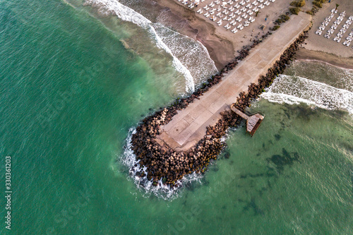 Aerial view of a concrete pier on sea shore with wave breaking stones.
