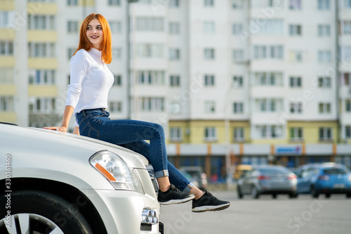 Young stylish redhead woman in white sweater, fashionable blue denim jeans pants and black sneakers sitting on a hood of new expensive car on city street enjoying warm summer day. © bilanol