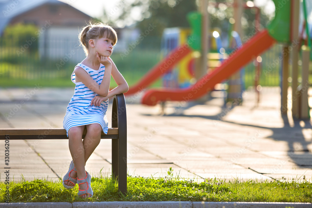 Cute young girl in short dress sitting alone outdoors on playground bench on sunny summer day.