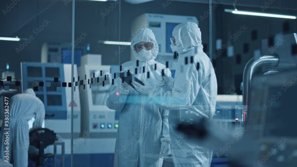 Research Factory Cleanroom: Engineer and Scientist Wearing Coveralls, Standing in Workshopop Talk and Use Tablet Computer, Professionals Develop Technology for Medical Electronic Equipment. Research Factory Cleanroom: Engineer and Scientist Wearing Coveralls, Standing in Workshopop Talk and Use Tablet Computer, Professionals Develop Technology for Medical Electronic Equipment.