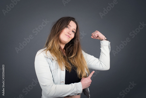 Waist up shot of blonde woman raises hand to show her muscles, feels confident in victory, looks strong and independent, smiles positively at camera, stands against gray background. Sport concept.