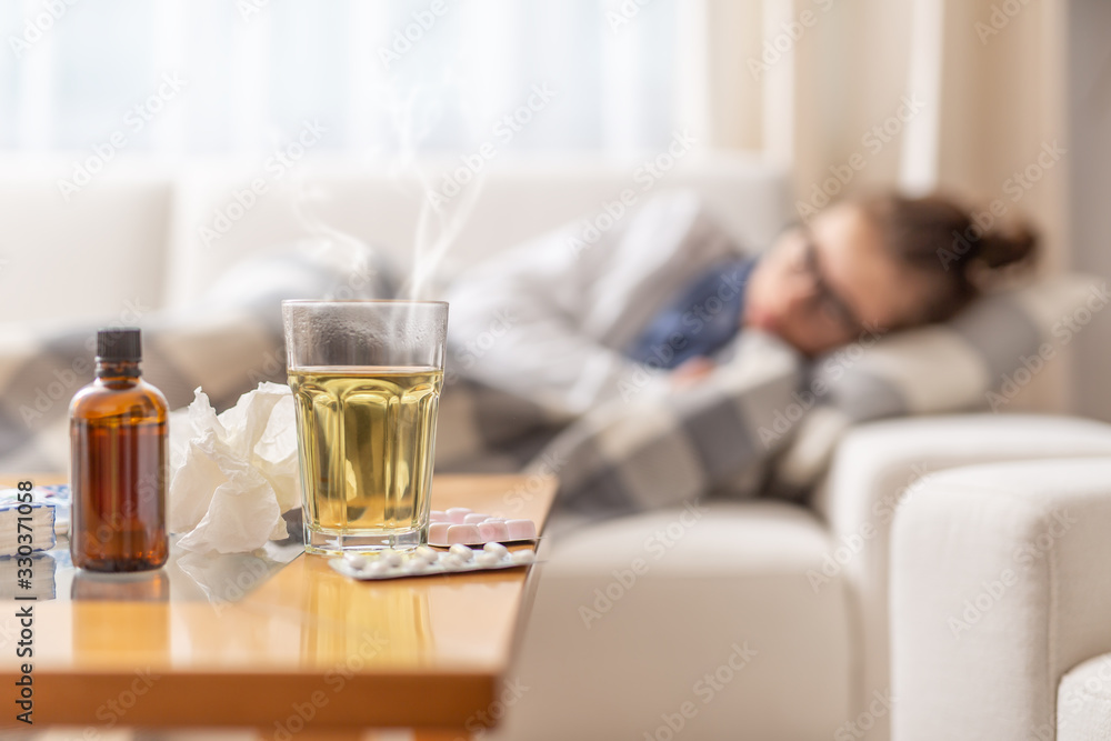Young ill woman lying under blanket on a bed with seasonal illnes drinking hot tea with honey and lemon. She takes pills and cough sirup to push the disease