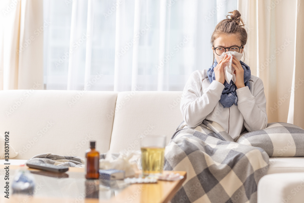Ill teen with runny nose sitting on a sofa at home and sniffling because of cold and fever. she has to use tissues and take pills and cough sirup