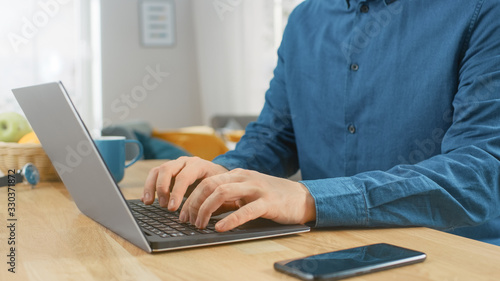 Close Up Shot of a Man Sitting at the Table at Home and Working on His Modern Silver Laptop. Smartphone Lies on a Wooden Table Next to Him.