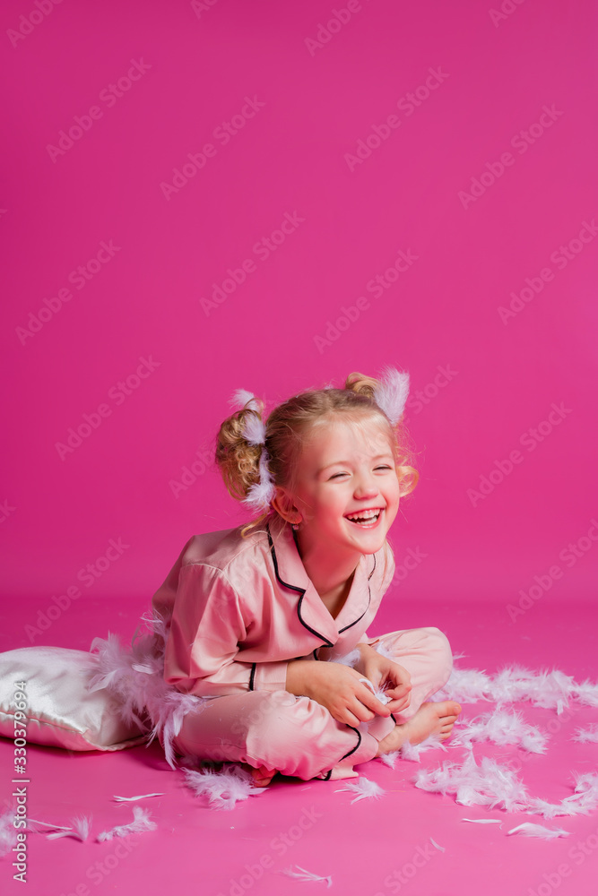 Studio shot of a contented, beautiful little girl posing in pink pajamas. Funny baby girl in pyjamas standing on a pink background with a pillow, feathers and a clock Studio shot of a contented, beautiful little girl posing in pink pajamas. Funny baby girl in pyjamas standing on a pink background with a pillow, feathers and a clock