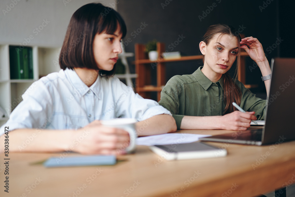 Two young woman working, looking at laptop screen, making notes and drinking coffee Two young woman working, looking at laptop screen, making notes and drinking coffee