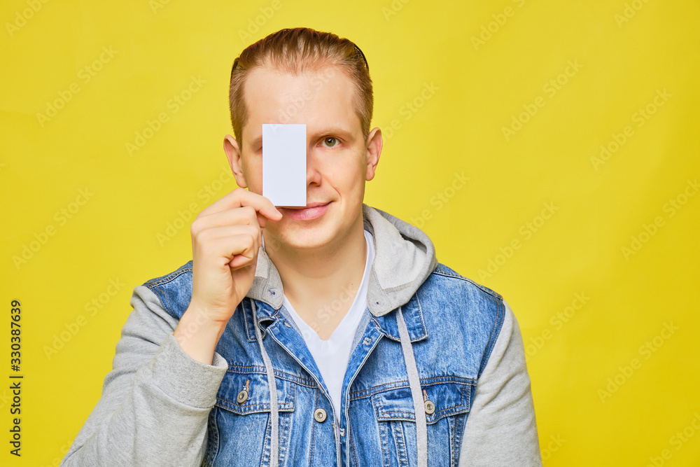 Stylish Caucasian man in a jeans on a yellow background holds a white card at eye level and smiles copy space. Stylish Caucasian man in a jeans on a yellow background holds a white card at eye level and smiles copy space.
