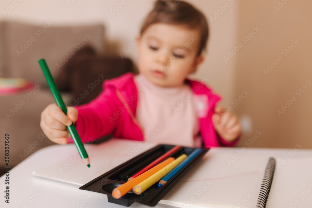 Happy baby girl sit at the table and wthire something. Little girl use pencil for drawing on white paper at home Happy baby girl sit at the table and wthire something. Little girl use pencil for drawing on white paper at home