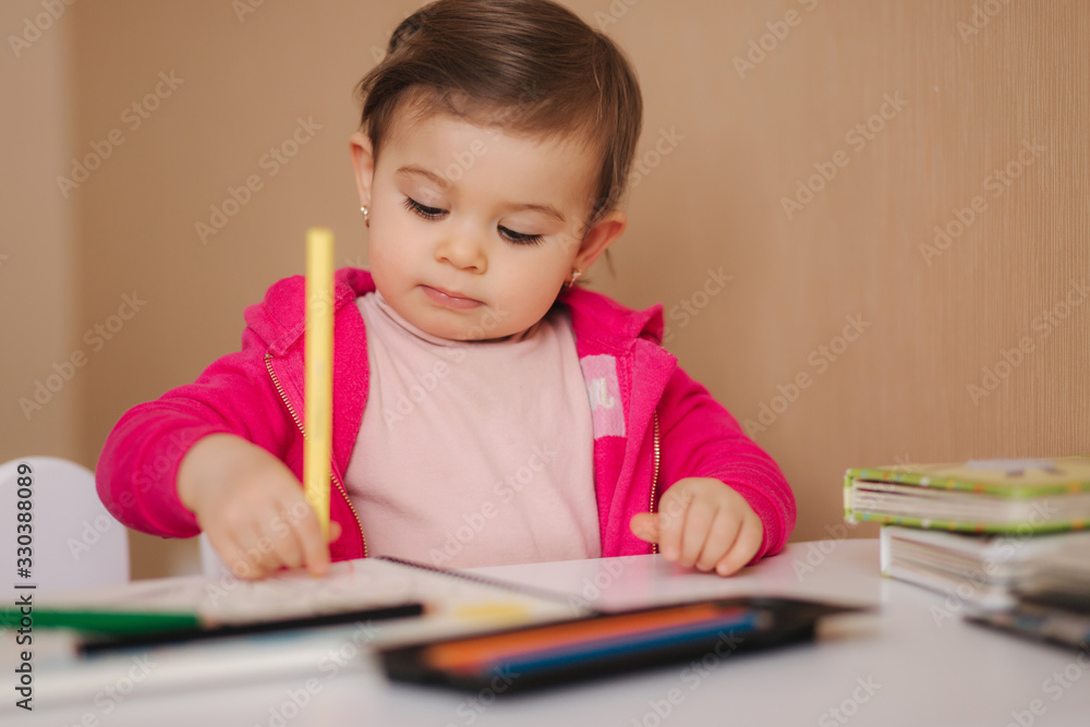 Happy baby girl sit at the table and wthire something. Little girl use pencil for drawing on white paper at home Happy baby girl sit at the table and wthire something. Little girl use pencil for drawing on white paper at home