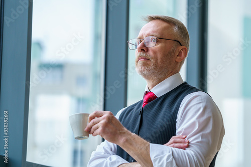 Businessman wears neatly white shirt. Man drinking coffee near big window. City view. Senior looking away.