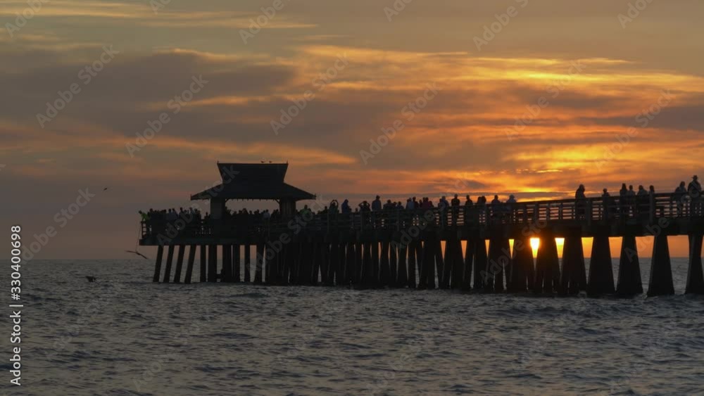 People walk along pier at sunset time. Sun sets in the ocean against backdrop of a pier in Naples, Florida. Naples Beach and Fishing Pier at Sunset. Dark silhouette of a pier over the water at sunset. People walk along pier at sunset time. Sun sets in the ocean against backdrop of a pier in Naples, Florida. Naples Beach and Fishing Pier at Sunset. Dark silhouette of a pier over the water at sunset.