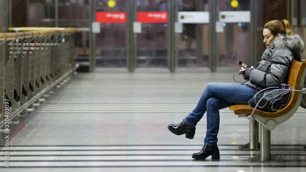 Young pretty lady sits on wooden bench in spacious lit lobby with phone in her hands and plugs in power bank. Successful woman in winter jacket with fur collar on platform of subway in Moscow, Russia Young pretty lady sits on wooden bench in spacious lit lobby with phone in her hands and plugs in power bank. Successful woman in winter jacket with fur collar on platform of subway in Moscow, Russia