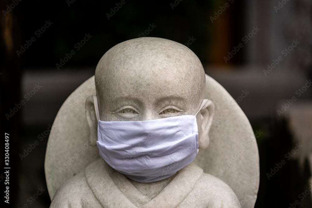 Portrait of buddhist statue wearing a face mask, closeup. Concept flu epidemic, coronavirus, dust allergy, protection against virus. White protective mask against coronavirus on a buddhist statue Portrait of buddhist statue wearing a face mask, closeup. Concept flu epidemic, coronavirus, dust allergy, protection against virus. White protective mask against coronavirus on a buddhist statue