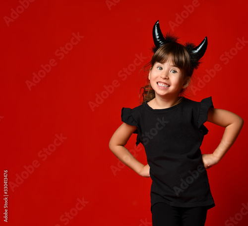 Little girl with devil horns, in black blouse and leggings. She laughing, hands on hips, posing on red background. Close up