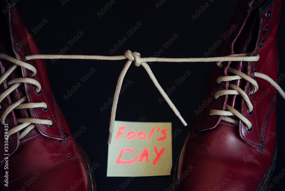 Stylish red shoes with laces tied together on black background. Boots covered by sticky note with text. April fool's day concept.