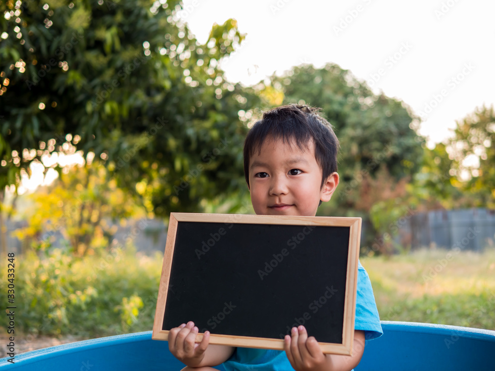 Fototapeta premium Asian cute little child boy holding empty blank black board with happy smiling face outdoor with green rural nature background. Young kid in blue t-shirt in family fun time. Copy space for education.