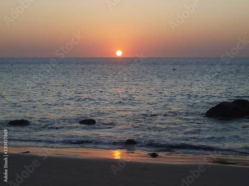 The sun moving over the horizon of the sea during sunset. In the foreground are sand  a mountain  stones and a water surface