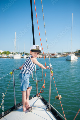 happy cute little caucasian boy in captain white hat striped tank top and denim shorts standing on luxury sea yacht in port during summer cruise travel