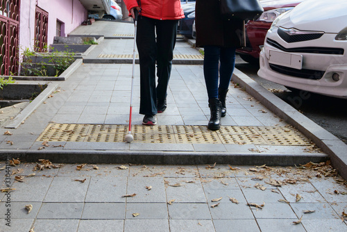 Blind person is walking on the sidewalk, using a white cane.