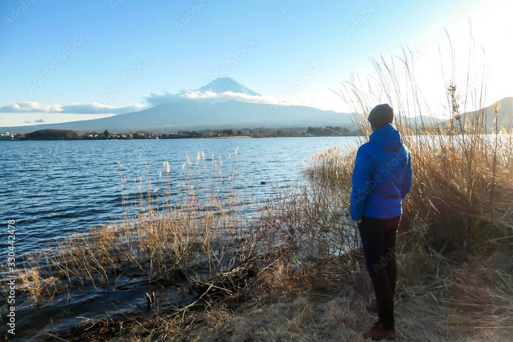 A man walking in between golden grass at the shore of Kawaguchiko Lake, Japan with the view on Mt Fuji. The man is enjoying the view on the volcano. The mountain surrounded by clouds. Serenity A man walking in between golden grass at the shore of Kawaguchiko Lake, Japan with the view on Mt Fuji. The man is enjoying the view on the volcano. The mountain surrounded by clouds. Serenity