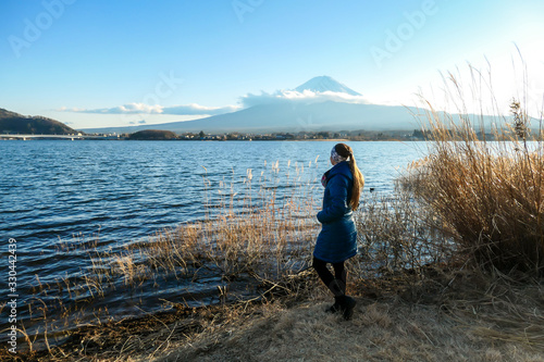 A woman walking in between golden grass at the shore of Kawaguchiko Lake, Japan with the view on Mt Fuji. The girl is enjoying the view on the volcano. The mountain surrounded by clouds. Serenity