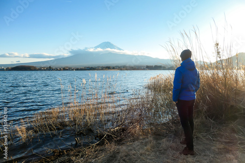 A man walking in between golden grass at the shore of Kawaguchiko Lake, Japan with the view on Mt Fuji. The man is enjoying the view on the volcano. The mountain surrounded by clouds. Serenity