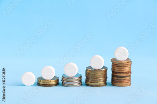 Growth graph made of stacked coins with white pills on the top of coins on the blue background. Growing market and increasing demand for medicine. Theme of the rise in price of health.