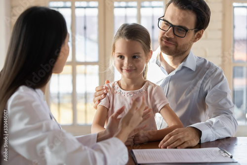 Front view young handsome father in eyeglasses holding on lap smiling cute little daughter, listening to pediatrician at meeting. Female doctor explaining vaccination details to interested patients.