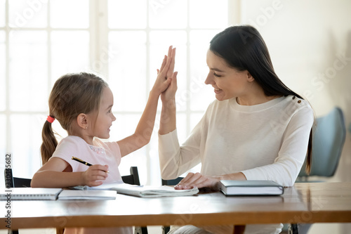 Joyful small daughter giving high five to satisfied with homework results mother. Happy young mom teacher babysitter praising little preschool child girl for making right tasks, sitting at home.