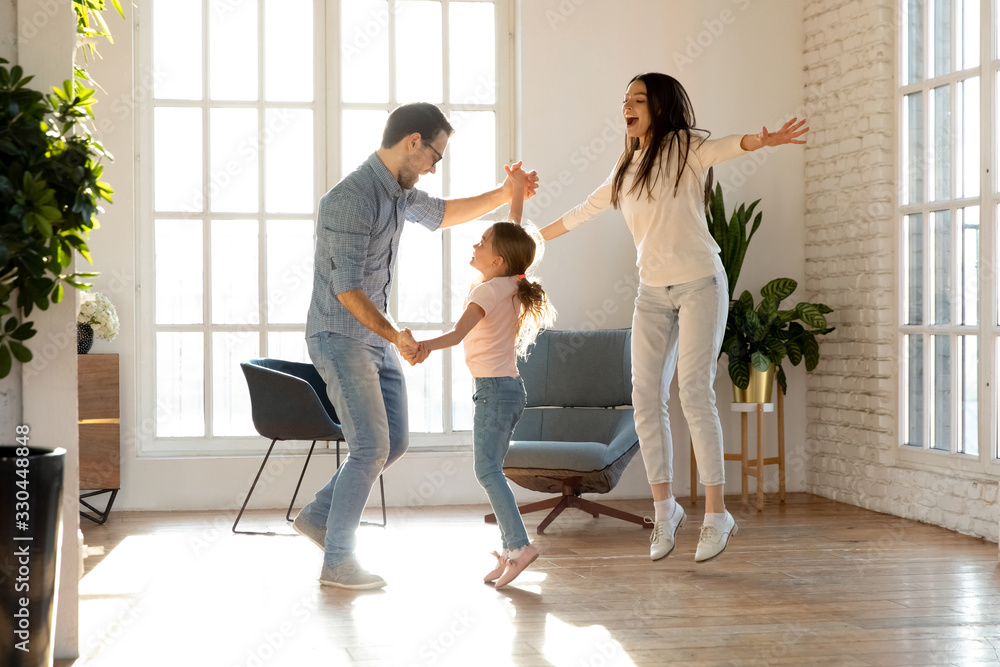 Full length overjoyed family of three having fun together. Smiling young father holding hands of happy little daughter, dancing together to favorite music while joyful mother jumping singing song. Full length overjoyed family of three having fun together. Smiling young father holding hands of happy little daughter, dancing together to favorite music while joyful mother jumping singing song.