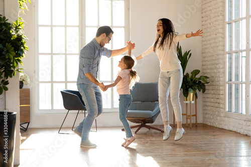 Full length overjoyed family of three having fun together. Smiling young father holding hands of happy little daughter, dancing together to favorite music while joyful mother jumping singing song.